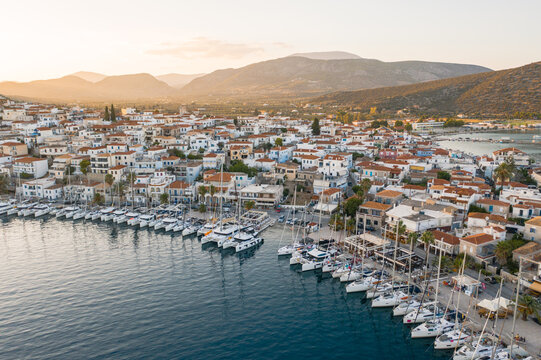 Aerial view of boats nestled in the harbor, buildings with terracotta roofs cascading towards the shimmering sea under a golden sky, Ermioni, Argolis, Greece.