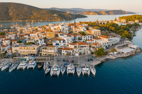 Aerial view of sun-kissed white buildings cascade towards the azure harbor where yachts bob gently, framed by rugged hills, Ermioni, Argolis, Greece.