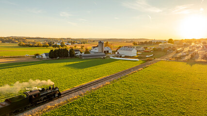 A vintage steam train chugs along railway tracks bordered by lush green fields. The sun sets over a...