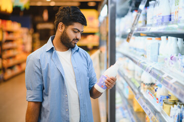 Young indian man choosing milk in supermarket refrigerator dairy section
