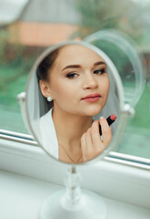 Bride applying lipstick in front of a mirror, her reflection highlights tenderness and elegance. Wedding preparation atmosphere, focusing on beauty, femininity, and graceful bridal style.