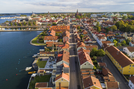 Aerial view of a vibrant town with red-roofed buildings nestled along the dark blue coastline, contrasting with the clear sky and lush greenery, Vastervik, Kalmar County, Sweden.