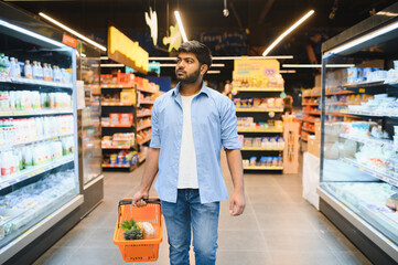 Young indian man walking through supermarket aisles holding shopping basket