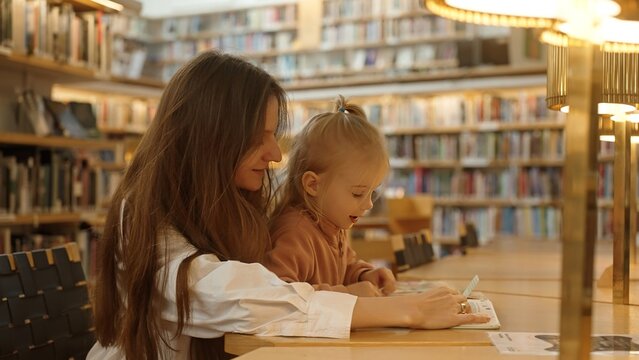 A young mom and her daughter enjoy reading a book at the library table. An adult parent or tutor assists with school preparation and homework. The concept of private ed for children. Finland Rovaniemi - Powered by Adobe