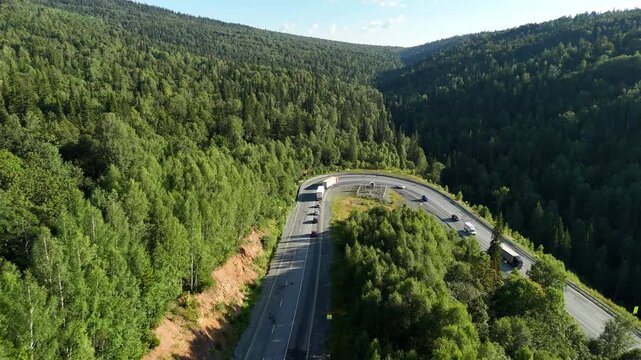 Aerial view of a sharp mountain highway curve with trucks and cars driving through dense green forest on a sunny summer day.