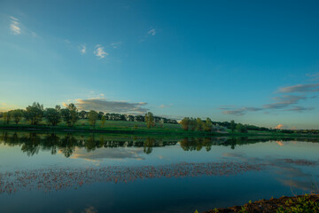 Obraz premium A wide horizontal view of a serene lake in the evening, with a clear blue sky and clouds reflected on the water, and a distant green treeline on the opposite shore