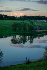 A picturesque rural scene at dusk with a calm lake reflecting the sky and trees. Pastel clouds are perfectly mirrored in the water, with a few distant houses visible beyond the treeline.