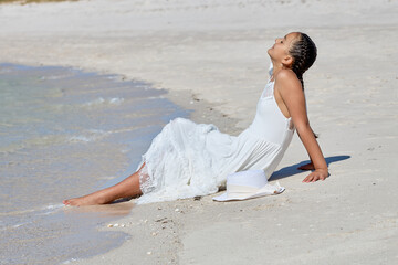 Young girl enjoys a sunny day on the beach while sitting by the water's edge in a flowing white...