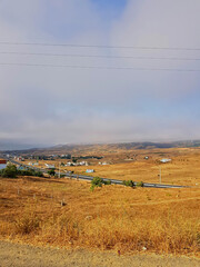 The overlapping meadows extend in warm light toward the horizon, where wavy hills gently meet the villages. A solitary asphalt road passes through the fields and houses.
