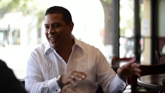 Charismatic Hispanic man in a white guayabera shirt smiles and gestures during a conversation in a cafe