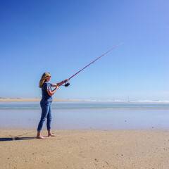 Barefoot woman on the seashore, holding a fishing rod, under a cloudless blue sky