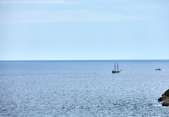 A traditional sailing ship cruises on the calm sea near a rocky coastline under a soft blue sky,...
