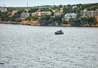 A modern motor yacht floats calmly in a coastal bay, with forested cliffs and residential buildings forming the scenic backdrop. leisure, architecture
