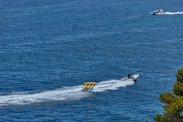 A speedboat pulls an inflatable banana ride with four people across clear blue sea on a sunny day,...