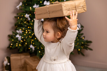Cute toddler girl in an elegant white dress stands in front of a blurred Christmas tree background,...