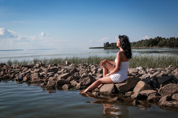 Young sexy beautiful woman is sitting on the rocks in the sea near the shore and enjoys relaxation, the warm sun and enjoys the sunset over the water. Wellness, tourism, holidays, vacation and weekend