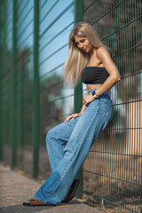 woman stands casually against a green fence, wearing a black crop top and wide leg blue jeans Her...