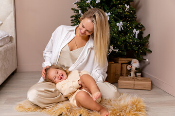 Mother and her little daughter sit on a floor under Christmas tree, happily spending time together,...