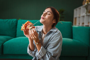Young woman enjoying pizza at home: licking finger with closed eyes