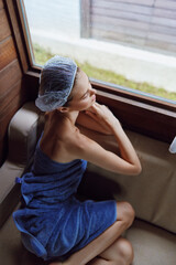 Relaxed woman with towel and shower cap sitting by window inside wooden sauna, enjoying peaceful...