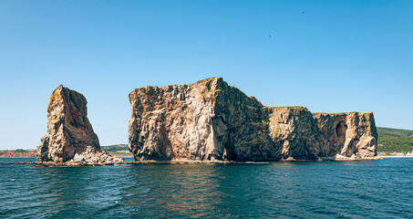 Large rocky cliff rising from the sea on a sunny day, seen from the water, with clear blue sky and waves crashing at the base.

