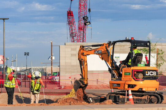 Construction crew operates JCB compact excavator to dig for sidewalk landscaping outside data center building site under construction, representing commercial development and utility work