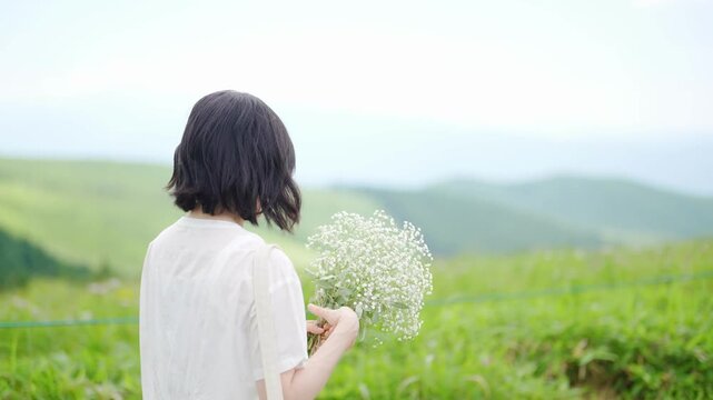 Slow-motion. In a wide, sunlit grass field under a bright blue sky, a Japanese woman in a flowing white dress walks away from the camera, holding a bouquet of baby's breath flowers. Nagano, Japan.