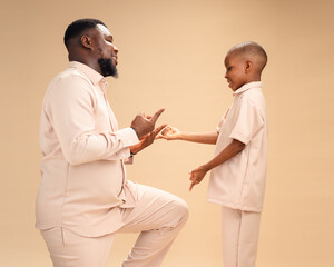 Man kneeling on one knee faces smiling boy in matching beige, both playfully mimicking a spider hand sign, radiating joyful father-son bonding, connection, and fun family moments.