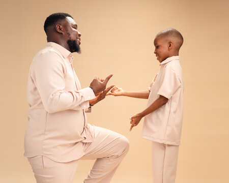 Man kneeling on one knee faces smiling boy in matching beige, both playfully mimicking a spider hand sign, radiating joyful father-son bonding, connection, and fun family moments.