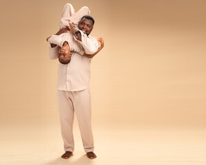 Under soft, warm light, a Nigerian father in beige playfully holds his son upside down. Their...