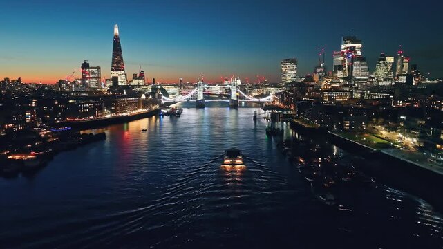Illuminated London city skyline at sunset, Tower Bridge, The Shard, and a ferry navigating the River Thames. Night lights cityscape. United Kingdom, Europe. Travel destination. Drone flight footage