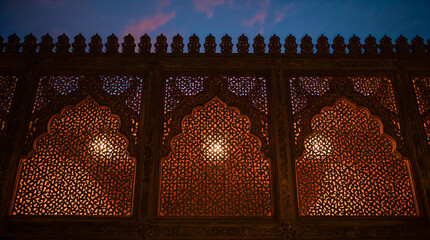Ornate wooden lattice screen with warm internal lighting against a dusky blue and pink sky, showcasing intricate geometric patterns and archways.
