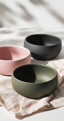 Three matte bowls, pastel pink, olive green, charcoal gray, on linen cloth, sunlight background, food serving