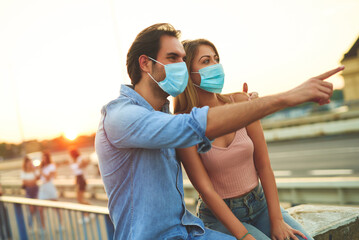 A young couple sits closely on a bridge, both wearing masks. They point towards the horizon,...