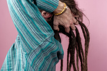 A rasta man is gently handling long dreadlocks while wearing a striped shirt. The pastel pink...