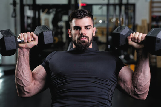 A man with a strong physique performs a bench press with dumbbells in a well-equipped gym. The atmosphere is energetic, emphasizing strength training and fitness