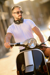 A handsome young man with sunglasses and headphones enjoys a ride on his scooter in an urban...