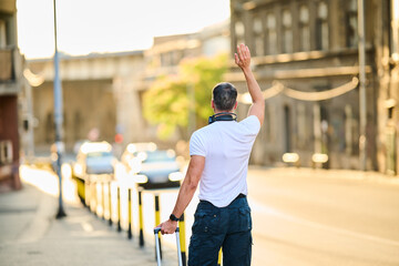 A man hailing a cab, trying to catch a taxi in an urban area, carrying luggage, holding his right...