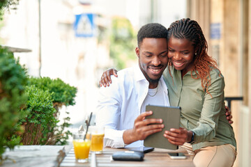 A young african american straight couple sits at a cafe terrace on a sunny day, smiling as they interact with a tablet. They are enjoying drinks while sharing joyful moments together