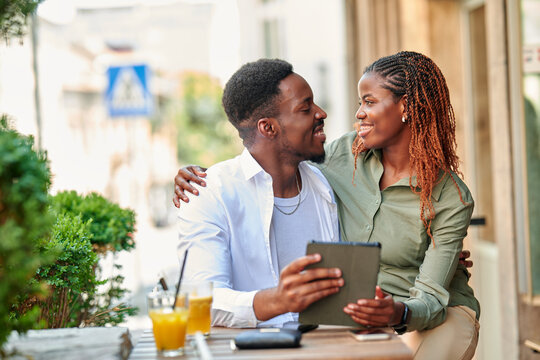 Straight african american couple sitting in a cafe, gazing at each other, holding a tablet and enjoying drinks
