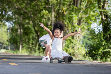 A joyful young girl spreads her arms wide while riding a skateboard on a forested path, smiling...