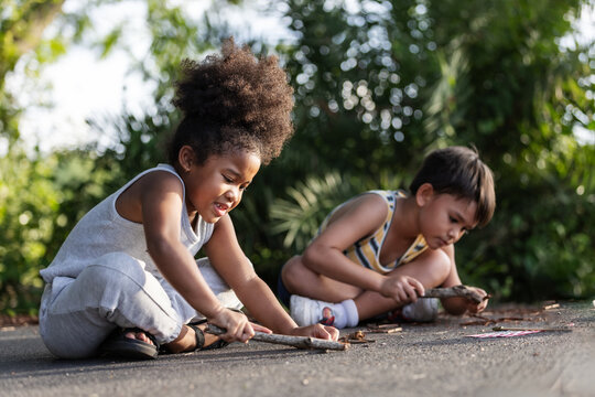 Two young children sit on the ground drawing with sticks, fully engaged in creative outdoor play. Surrounded by greenery, they express imagination, focus, and the joy of hands-on exploration.