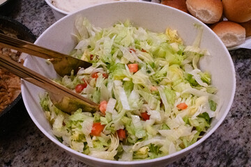 A fresh salad with shredded lettuce and diced tomatoes, served in a large bowl with golden serving tongs, accompanied by soft dinner rolls