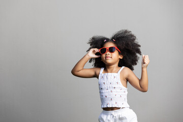A confident young girl in red sunglasses strikes a bold pose with both arms raised, exuding...