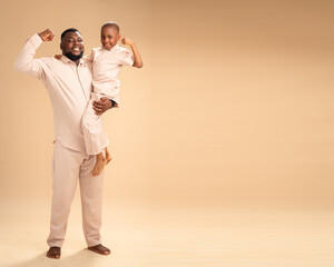 Father and son strike proud flexing poses in matching beige, set against a warm backdrop—a...