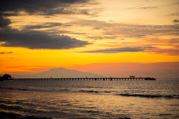 Obraz premium The beautiful orange-golden sunset sky at dusk on the coast of Dili, Timor-Leste. The silhouettes of the jetty and mountains can be seen in the distance.