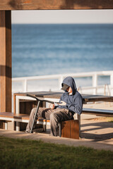 Boy sitting alone with scooter by the beach looking down at phone
