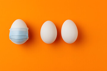 White egg wearing protective medical face mask beside two plain eggs on bright orange background, concept of food safety health protection pandemic coronavirus prevention hygiene creative still life


