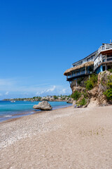 Fototapeta premium Praia tropical em Curaçao com águas azul-turquesa, areia branca e turistas aproveitando o dia ensolarado, cercada por natureza e falésias rochosas.