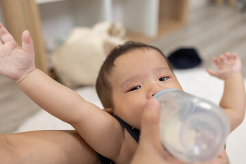 The baby is drinking milk from a bottle. The baby is being held by a person. The baby is wearing a blue shirt. This picture shows the care of the baby.
